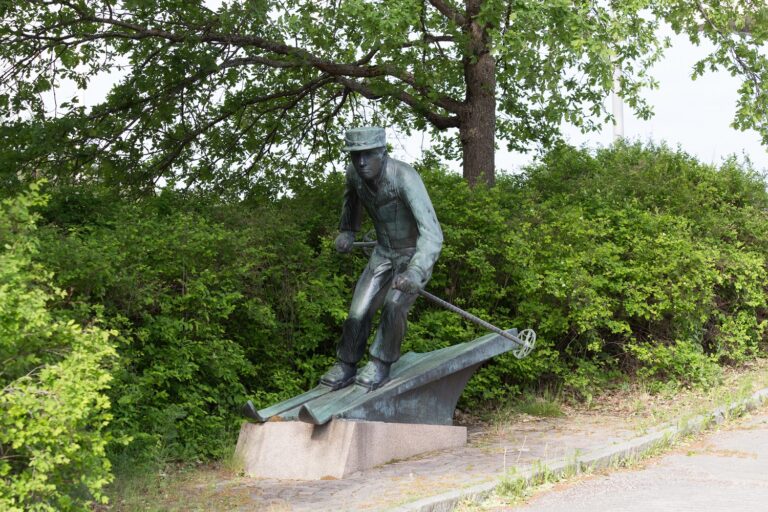 A bronze sculpture depicting a man in a classic skiing attire gliding downhill. He is bent forward, with arms and ski poles pointing outwards.