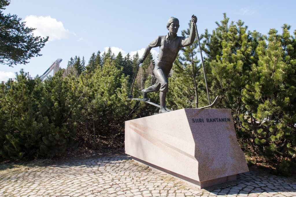 A bronze statue of a skiing woman. A ski jumping tower can be seen in the background behind the statue.