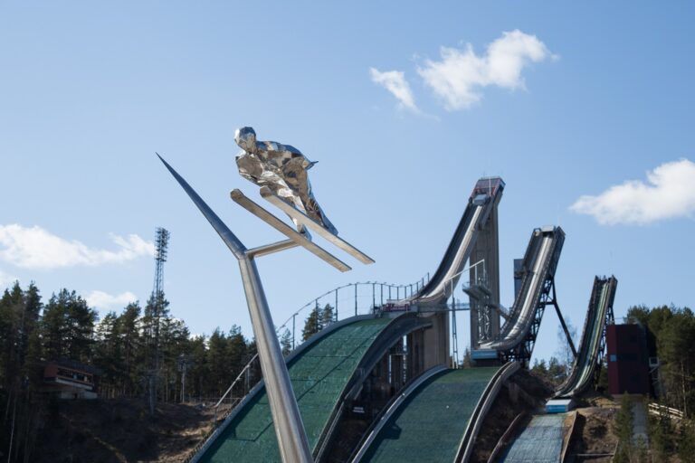 Steel sculpture of a ski jumper. The ski jumper is supported by a high steel boom. Stadium in the background.