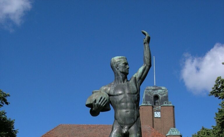 A bronze sculpture depicting a man posing naked, holding a helmet in one hand and the other hand up in the air.