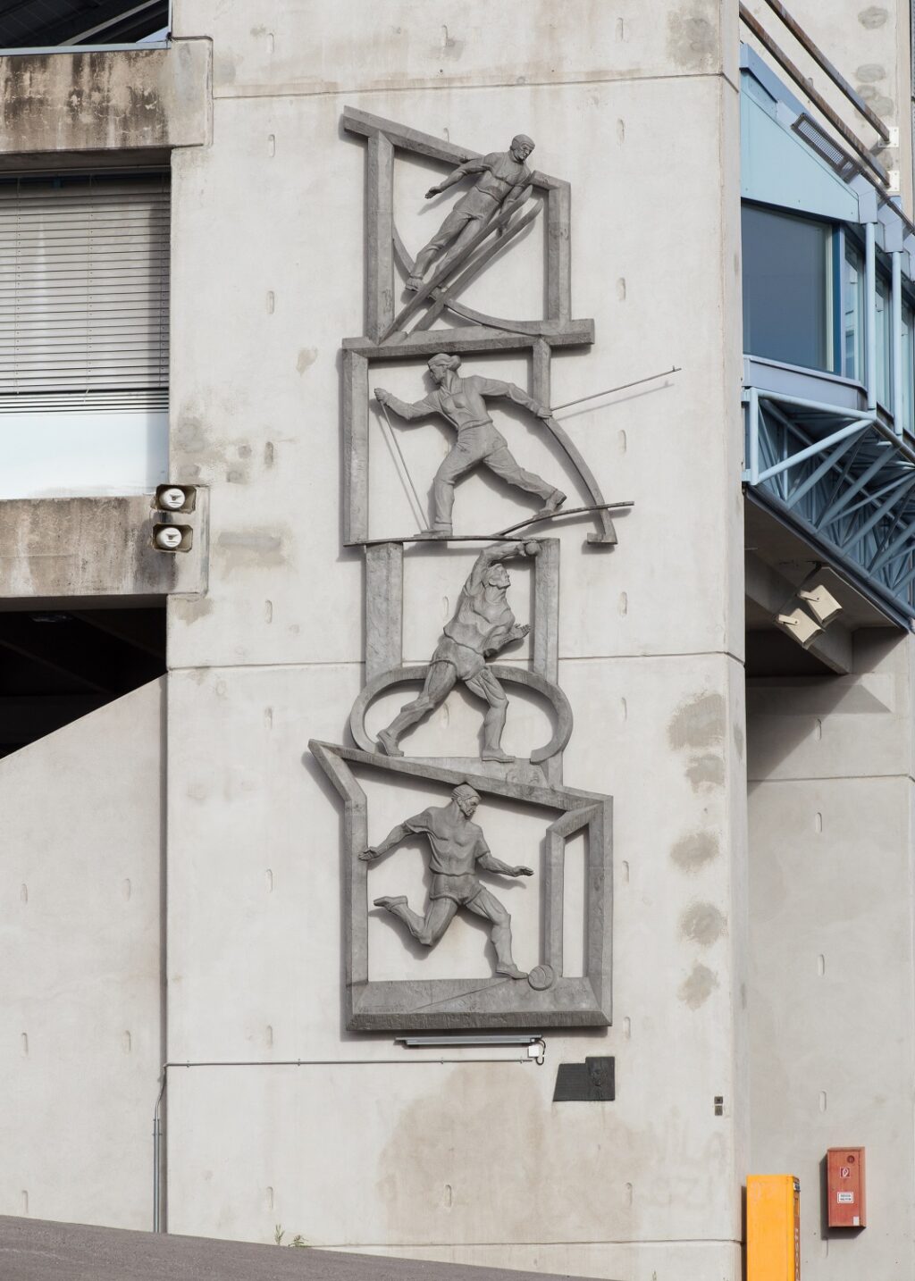 A realief attached to the outer wall depicting four different sports events on four “levels”: on top is a ski jumper, under him a female skier, then a shot-putter and at bottom a running football player.