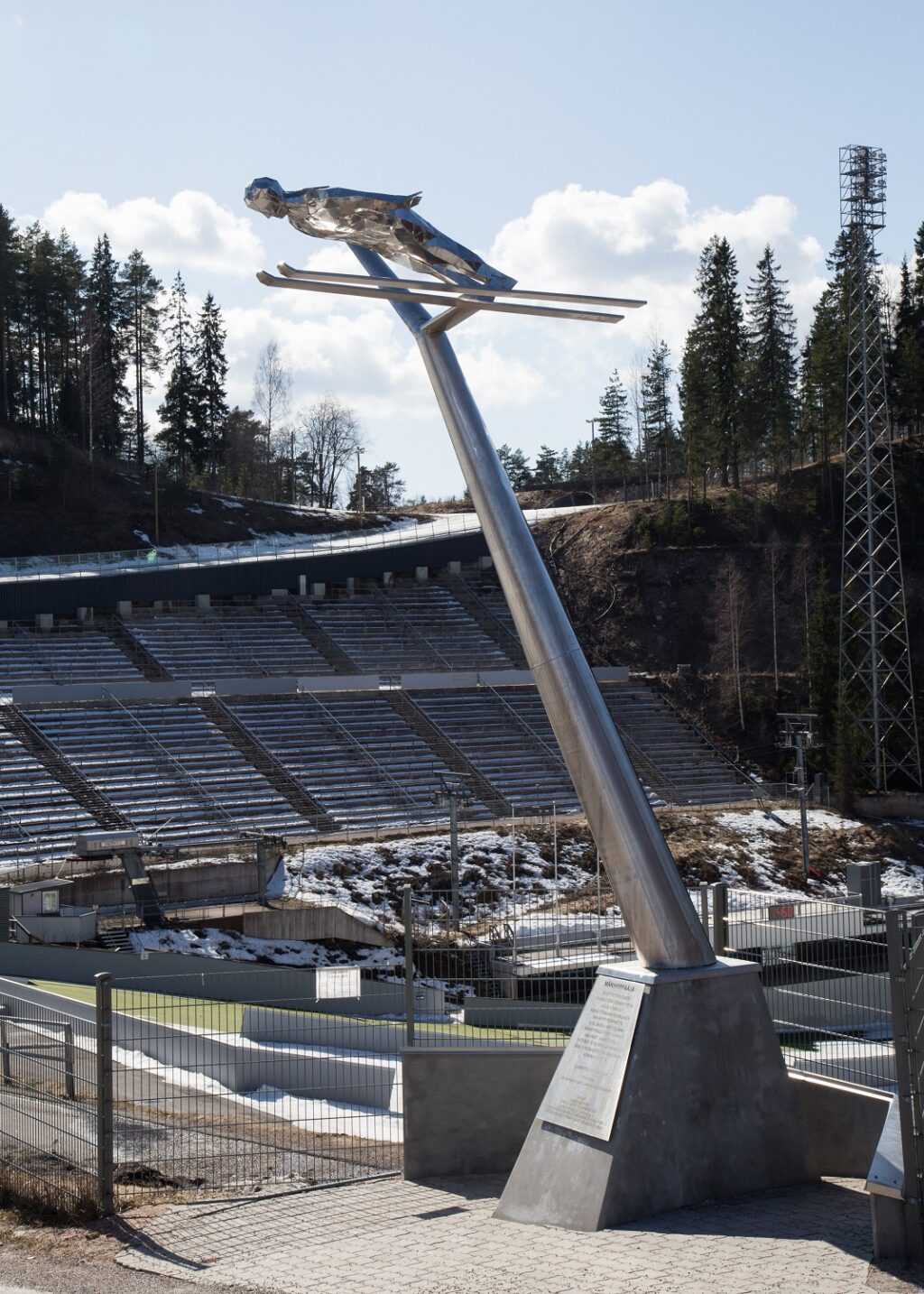 Steel sculpture of a ski jumper. The ski jumper is supported by a high steel boom. Stadium and grandstand in the background.