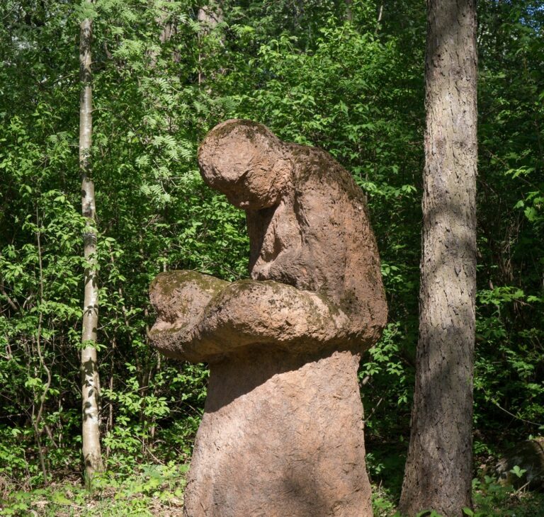 A concrete sculpture placed in the park depicting a human figure sitting on an anthill with their legs crossed.