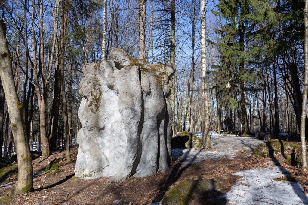 A concrete sculpture placed in a park depicting a crowd of standing nude figures gathered together.