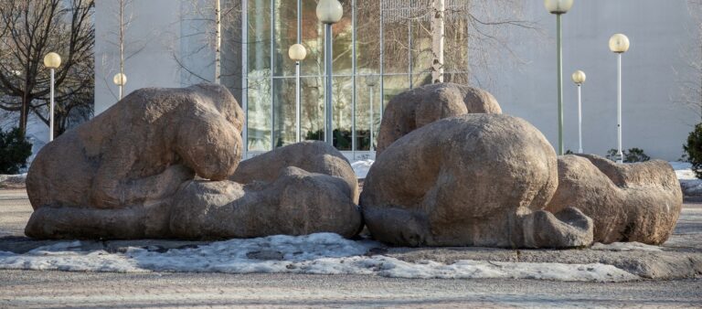 A set of concrete sculptures placed in front of the building, depicting five human figures in different positions against the ground.