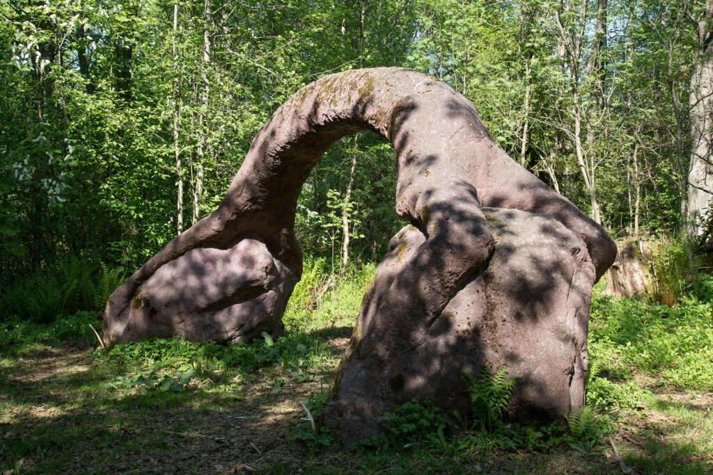 Puistoon sijoitettu betoninen veistos, joka kuvaa ihmishahmoa, joka on aseuttunut kahden kiven päälle kaarimaiseen, siltaa muistuttavaan asentoon.