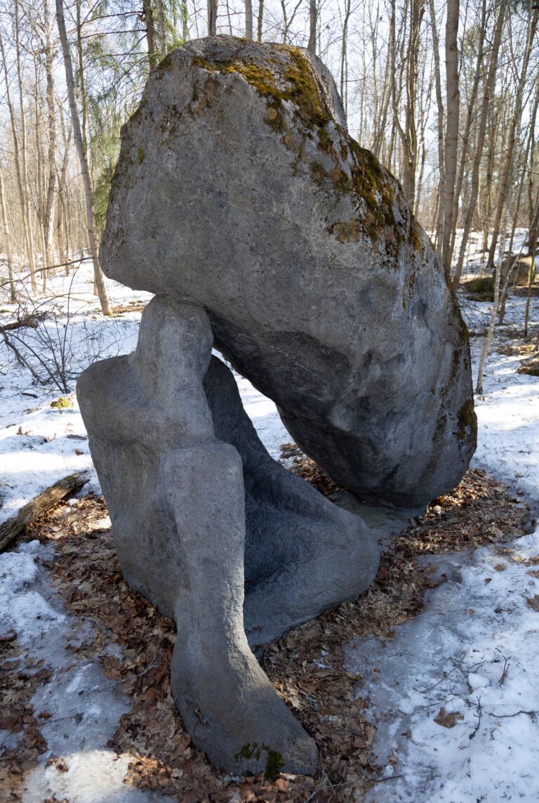 A concrete sculpture placed in the park, which consists of two interconnected parts: a large slanting stone pillar and a human figure holding it on its head.