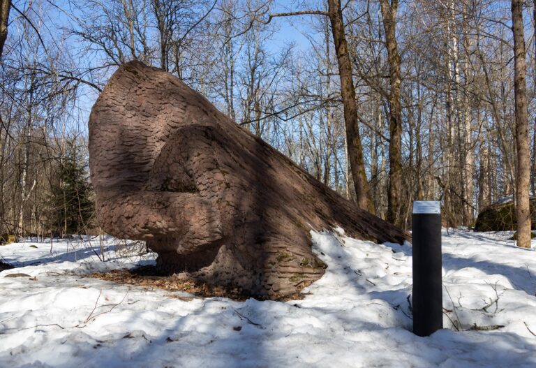 A concrete sculpture placed in the park, depicting a pile of small tree trunks trimmed of their branches.