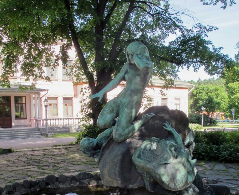 A bronze fountain sculpture depicting a nude female figure sitting on a stone with two human figures reaching under the stone towards her.