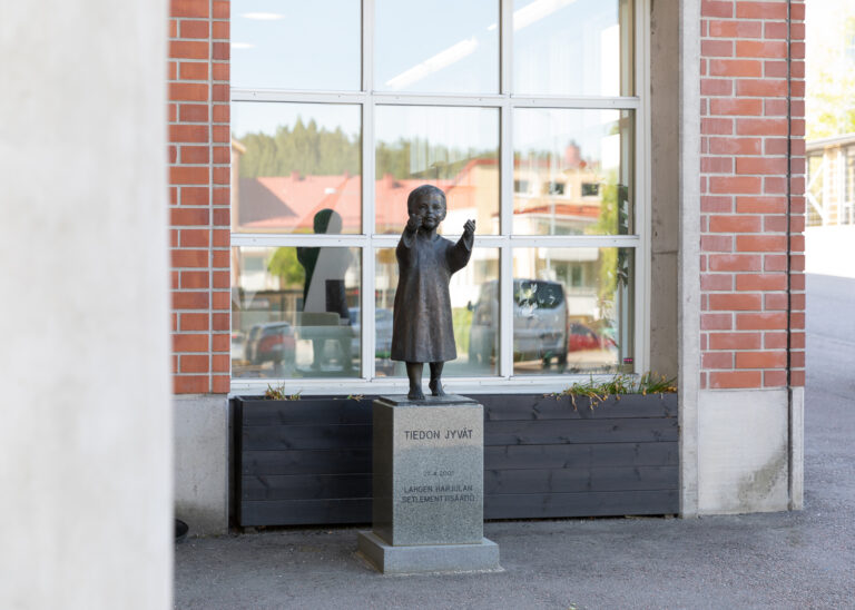 A bronze statue placed in front of a building, depicting a barefoot child dressed in a long dress holding out their hands from which grains fall.