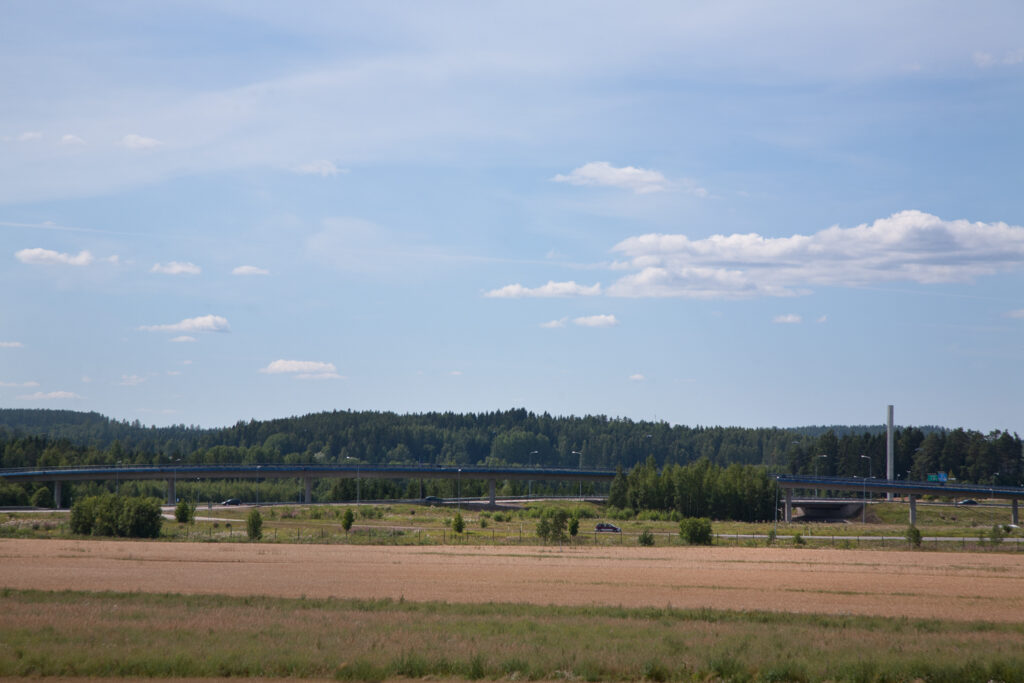 Peltomaisema, jonka taustalla näkyy siltaramppi, jonka kaide hehkuu sinisenä.