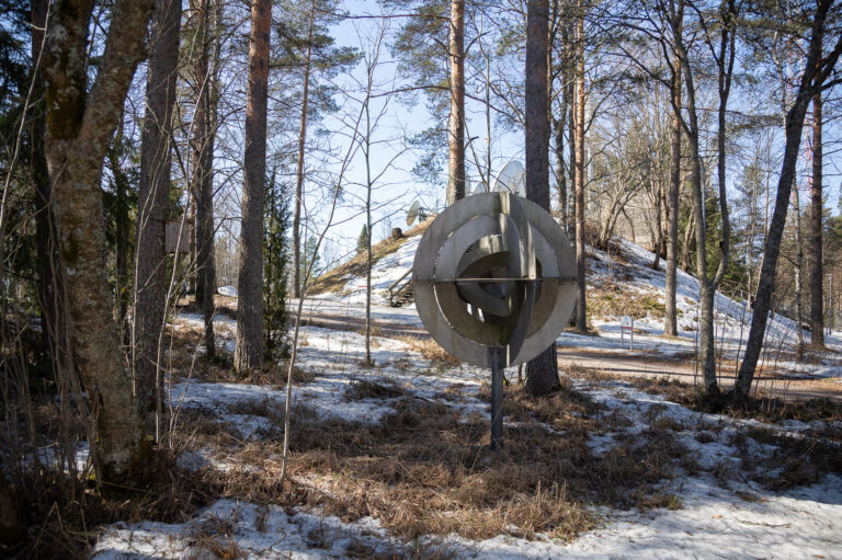 A steel, abstract sculpture resembling a satellite dish placed in the forest.