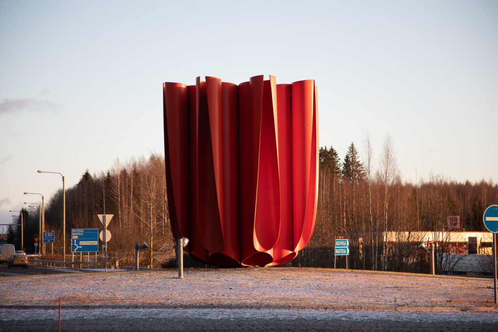 A plastic sculpture placed in a traffic circle. The sculpture is made of plastic water pipes painted bright red, which form the shape of a crucible used to melt metal.