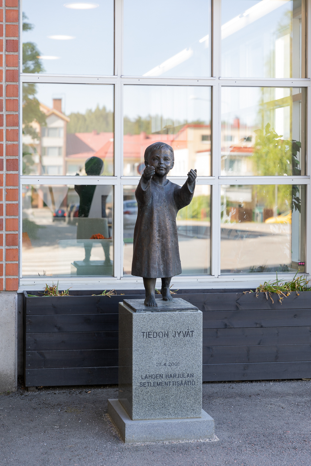 A bronze statue placed in front of a building, depicting a barefoot child dressed in a long dress holding out their hands from which grains fall.