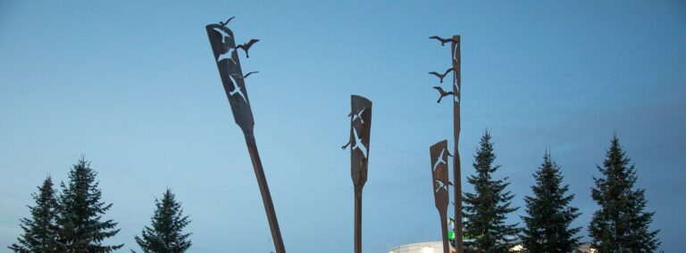 A metal sculpture with four oar-like parts rising from the ground. There are holes cut into their blades that form the silhouettes of birds. The actual iron bird figures cut from the pallets are attached to the wingtips of the silhouette birds.