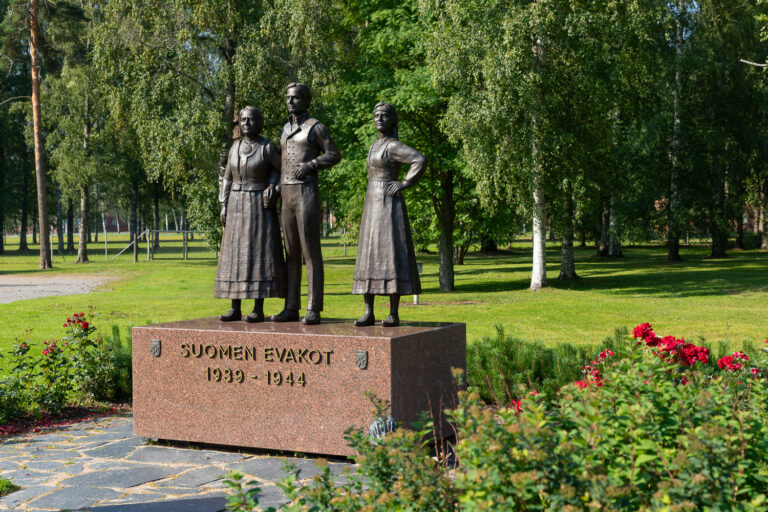 Statue of three bronze human figures on top of a square granite pedestal, captured slightly from the side, behind a red-toned rose bush.