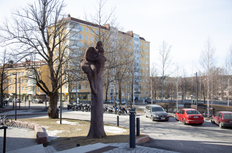 A concrete sculpture placed in the schoolyard, depicting a tree. At the top of the tree, there are two human figures sitting together.