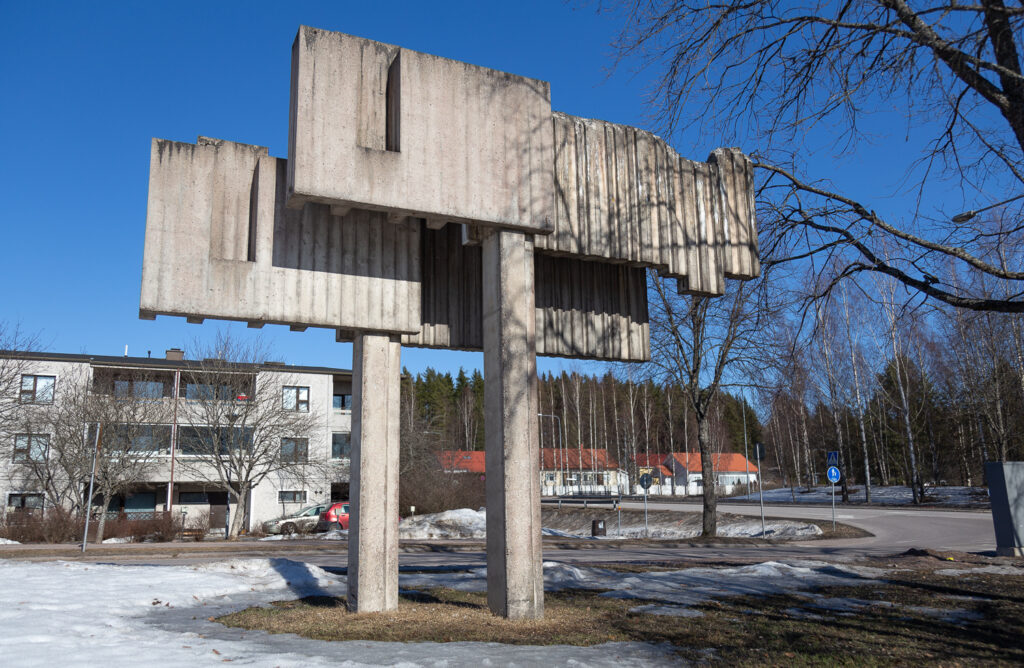 A concrete monument at the road intersection. In the background, there is an apartment building and further away, some detached houses.