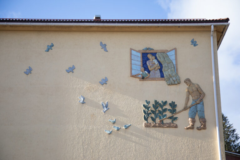 A ceramic relief attached to the wall of the building, depicting a man working in the field while the rest of the family watches flying birds in the sky through the window.