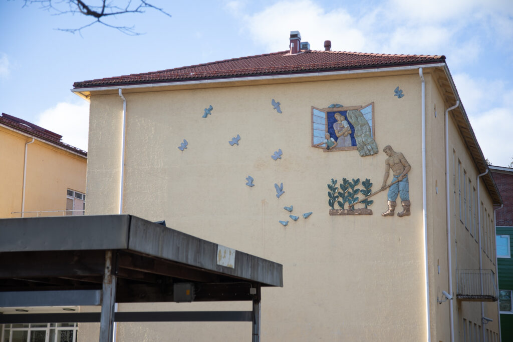 A ceramic relief attached to the wall of the building, depicting a man working in the field while the rest of the family watches flying birds in the sky through the window.