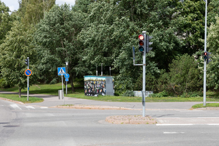 Air quality monitoring booth with a colorful mural at the roadside intersection. The intersection is lined with various traffic signs, and the traffic lights for both cars and pedestrians are red.