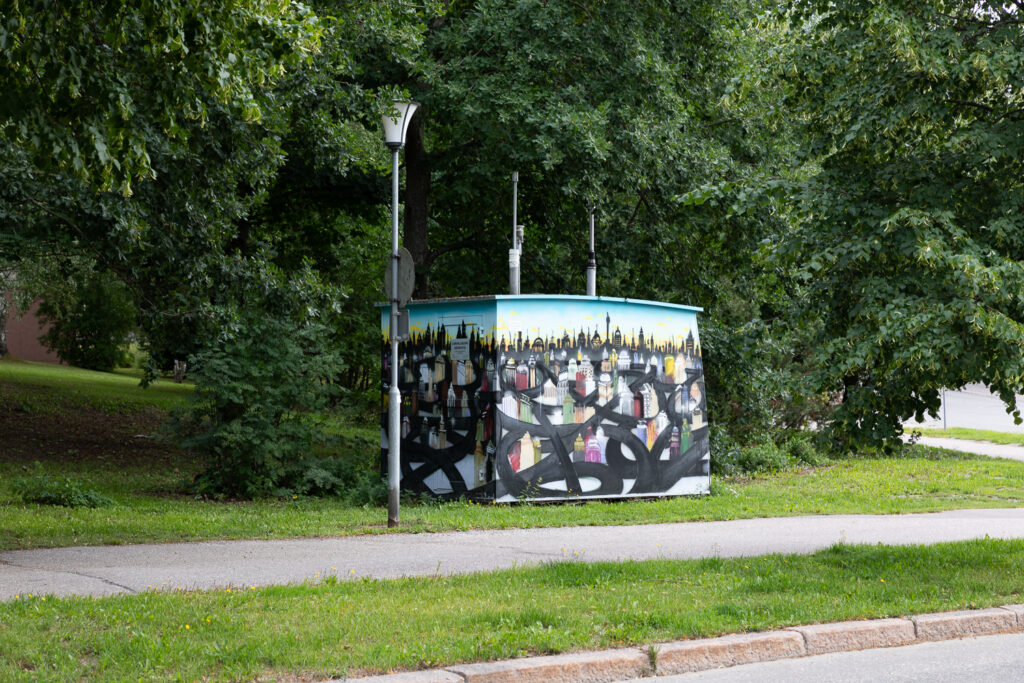 Air quality monitoring booth along a pedestrian path. The booth's roof and right side are curved with antennas on the roof. The booth features a colorful cityscape mural.