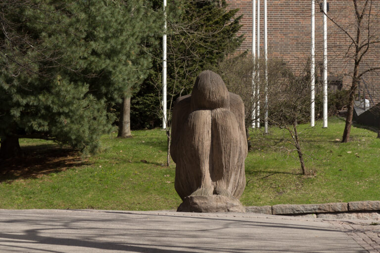 A concrete sculpture placed on the street depicting a seated human figure leaning on their knees.