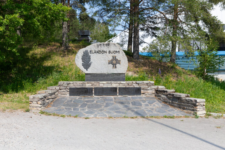 A concrete monument placed on top of a wall-like structure. On the monument, on the left, there is an oak leaf symbol, in the center the text reads 'Long Live Finland,' and on the right, the Mannerheim Cross. Behind the monument, a lush hill rises on the left, and in the background on the right, there is a large hall.