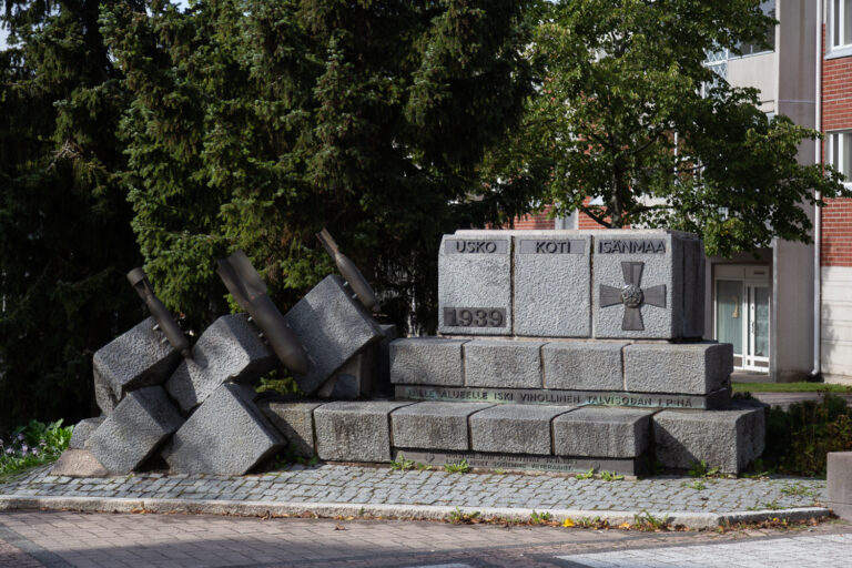 A slab placed on top of a stepped stone plinth, on which the words "FAITH, HOME, MOTHERLAND" are written on bronze plates attached to it. A composition depicting falling bombs and stones is grouped on the left side.