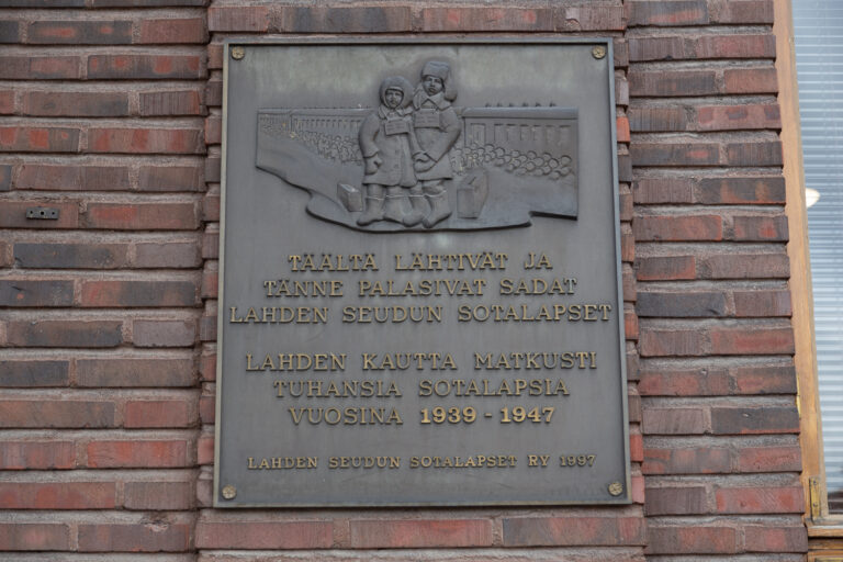 Relief attached to the red brick building's wall, depicting two children with their bags in front of a train, along with accompanying text.