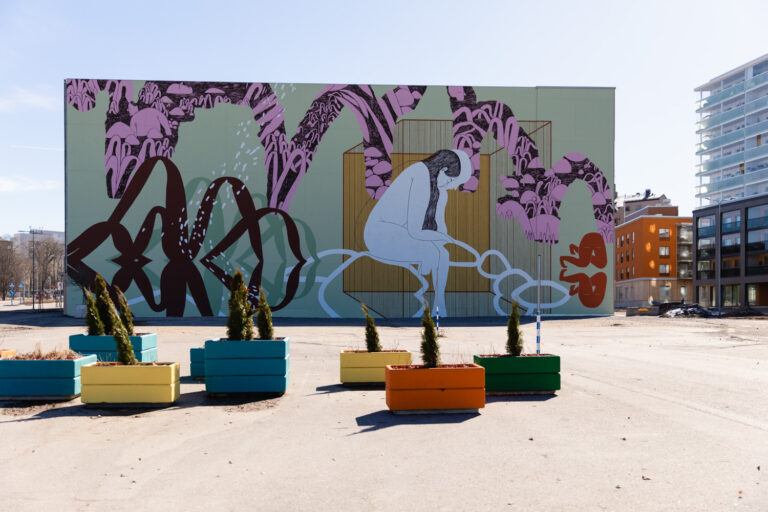 A huge, colorful mural on a rectangular shaped multistory car park building. There are plant boxes in many colors in the foreground of the image.