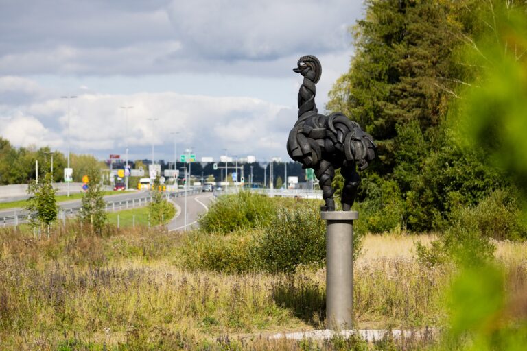 A sculpture depicting an ostrich made of car tires standing on a stand by the side of the road.